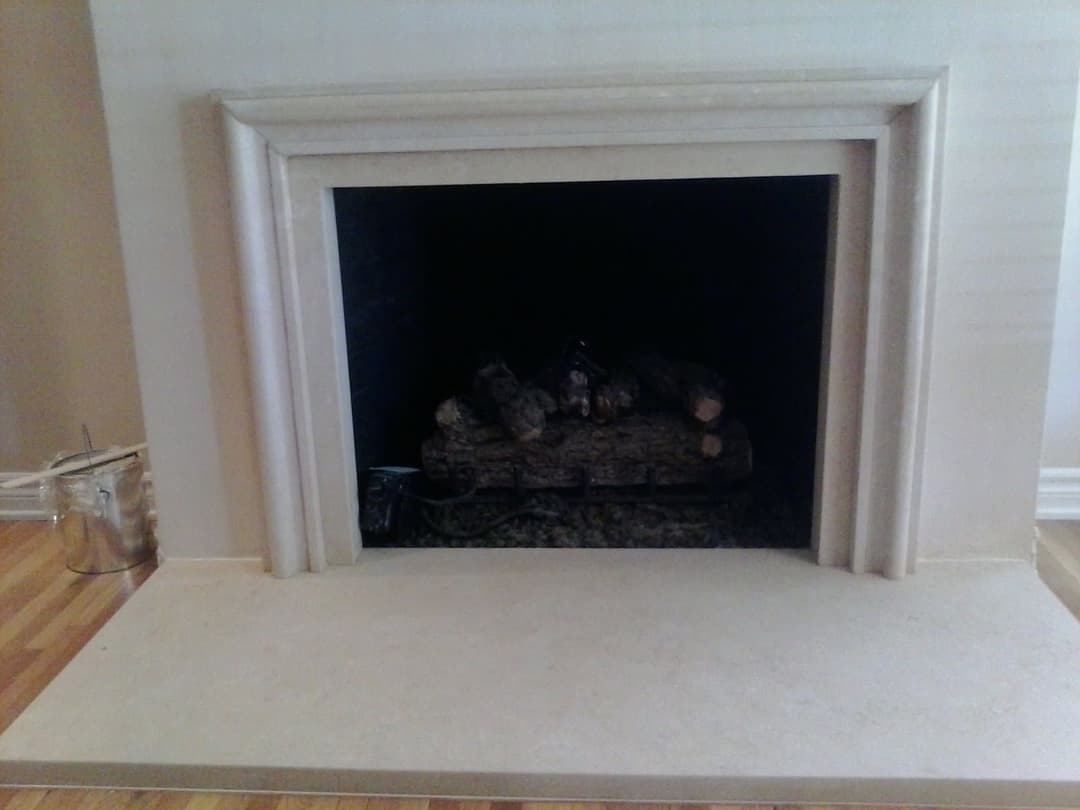 Empty fireplace with logs on the grate, framed in a cream-colored stone surround.