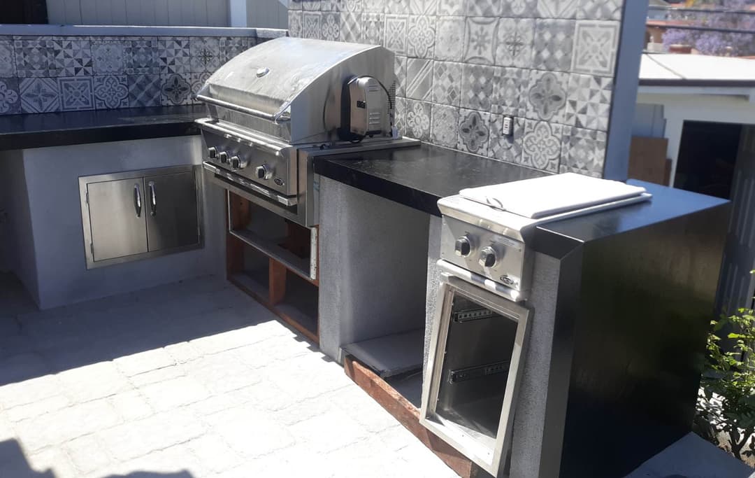 Outdoor kitchen with modern stainless steel grill, cooking appliances, and decorative tile backsplash.